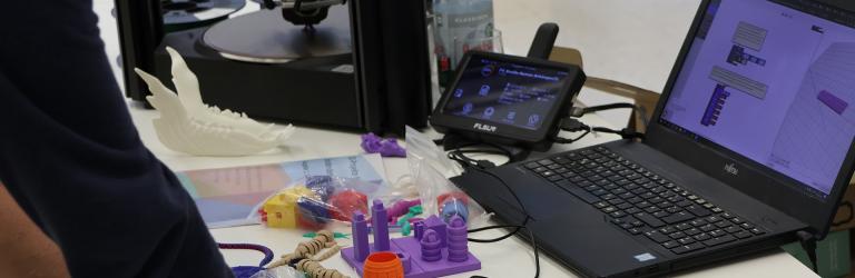  Hände stützen sich auf einen Tisch mit aufgeklapptem Laptop, dessen Screen ein 3D-Druck-Programm zeigt, Davor liegen bunte mit dem 3D-Drucker ausgedruckte Objekte. Links ist der 3D-Drucker zu erkennen.