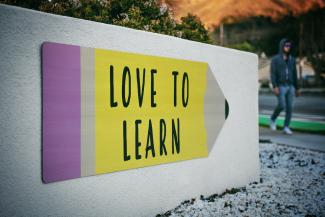 Auf einer Mauer ist ein großer Buntstift abgebildet, der den Schriftzug LOVE TO LEARN trägt und nach rechts auf einen Passanten weist