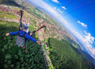 Junge Person (von hinten aufgenommen) schwebt im Tandem-Paragliding-Flug über Wald- und Stadtlandschaft, die Arme ausgebreitet