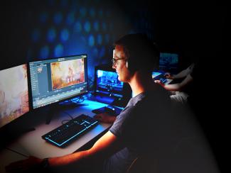 In einem angedunkelten Raum sitzt ein junger Mann vor 2 großen Screens, auf denen das Videospiel „Bioshock Infinite“ und eine Videoschnittsoftware zu sehen sind; im Hintergrund ist eine zweite Person erkennbar.