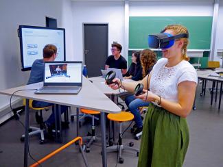 Innenansicht des neuen Lab-Standorts P18: Neben einem dreieckigen Stehpult nutzt eine Studentin das VR-Set; im Hintergrund sind vor einem Smartcreen weitere Personen um einen flexiblen Arbeitsplatz gruppiert.