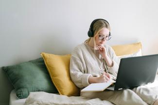 Eine Studentin sitzt mit Laptop und Kopfhörern auf dem Sofa und macht sich Notizen auf einem Block