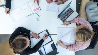 Blick von oben auf 2 junge Frauen, die mit Laptop und anderen Arbeitsmaterialien am Schreibtisch sitzen. Am oberen Bildrand sind die Hände weiterer Personen erkennbar.