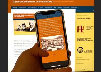 Vor dem Hintergrund der Website zur Ausstellung „Schliemann und Heidelberg“ hält eine Hand ein Smartphone, auf dessen Screen die digitale Schnitzeljagd via Actionbound zu sehen ist.