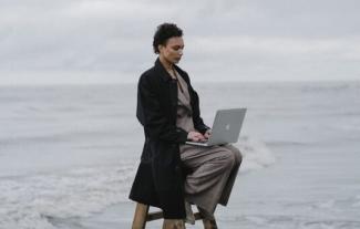 Vor winterlicher Meereskulisse sitzt eine junge Frau im schwarzen Mantel auf einer aufgeklappten Leiter in der Brandung. Auf ihrem Schoß ein aufgeklappter Laptop.