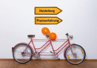 Ein orangefarbenes Tandem lehnt an einer Wand, am hinteren Lenker sind 2 orangefarbene Luftballons angebunden. Darüber der Schriftzug „Lehr:werkstatt“ in Orange, außerdem 2 Wegweiser: Auf dem einen steht „Heidelberg“, auf dem anderen „Praxiserfahrung“. 