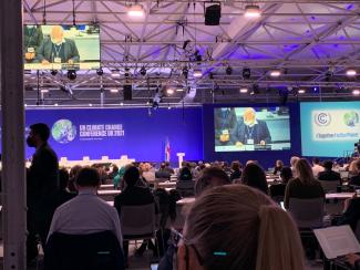 Szene auf der UN-Klimakonferenz in Glasgow 2021 (Conference of the Parties – COP26): Über die Köpfe vieler Teilnehmender hinweg sind große Screens zu sehen, die Tagungstitel und -Logo sowie einen Sprecher der EU zeigen. | © Max Jungmann