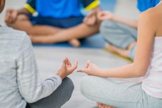 Junge Menschen verschiedenen Geschlechts und unterschiedlicher Hautfarbe sitzen zur Meditation in einem Kreis zusammen, die Hände in Mudras. Die Köpfe sind nicht zu sehen. 