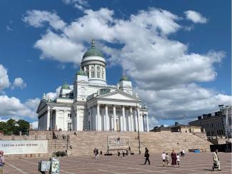 Man sieht den Dom von Helsinki bei schönem Wetter. Auf der großen Treppe und dem Platz davor spazieren einige Menschen.