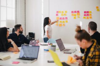 Studierende mit Laptops in einem modernen Seminarraum. Am Whiteboard eine Dozentin mit bunten Post-its.