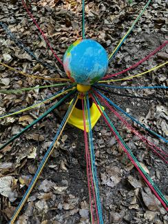 Eine Erdkugel steht auf dem Waldboden; strahlenförmig führen bunte Schnüre von ihr weg.