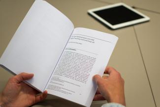 Zwei Hände haben den Sammelband aufgeschlagen; zu erkennen ist der Artikel zum HSE Digital Teaching and Learning Lab P18 von Bernd Hirsch und Timo Holste. Hinten auf dem Tisch liegt ein Tablet.