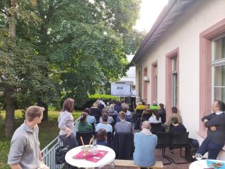 Das Bild zeigt die gut besuchte Terrasse der HSE; im Hintergrund der Referent vor einem Smartboard, links Bäume.