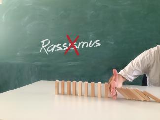 Auf einem Tisch sind in Domino-Art Holzklötzchen in Reihe aufgestellt; eine Menschenhand bremst den Dominoeffekt aus. Im Hintergrund eine grüne Tafel, auf der rot durchgestrichen das Wort „Rassismus“ steht.