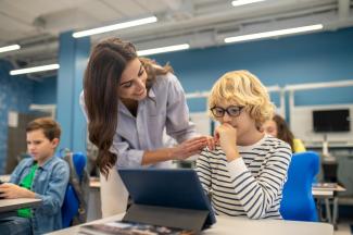 Eine Lehrerin beugt sich lächelnd zu einem Schüler, der nachdenklich vor einem Tablet sitzt. Im Hintergrund weitere Schüler:innen.