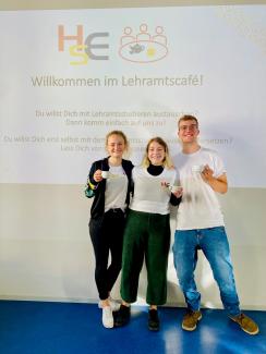 Drei lachende Lehramtsstudierende in HSE-Shirts vor dem Willkommensscreen des Lehramts-Cafés, in der Hand eine Tasse.