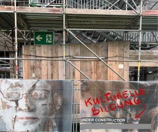 Das Bild zeigt ein eingerüstetes Gebäude, davor ein Bauzaun, der mit dem Plakat einer zerbrochenen Büste und dem Hinweis "UNDER CONSTRUCTION" versehen ist. Darüber in Rot: KULTURELLE BILDUNG: