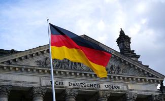 Das Bild zeigt den oberen Teil des Reichstagsgebäudes mit der Inschrift DEM DEUTSCHEN VOLKE, davor eine Deutschlandfahne.