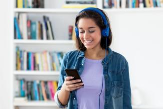 Das Bild zeigt eine lachende junge Frau mit Kopfhörern und Smartphone vor ihrem Bücherregal.