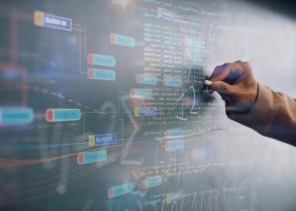 An einer Tafel, die mit blockbasiertem Code überzogen ist, notiert eine Hand mit Kreide mathematische Formeln.