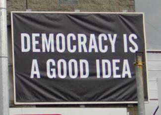 Auf einem schwarzen Banner steht in weißen Lettern DEMOCRACY IS A GOOD IDEA.