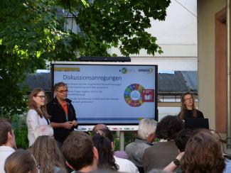 Teresa Ruckelshauß, Prof. Dr. Alexander Siegmund und Dr. Johanna Weselek vor einem Smartboard auf der Terrasse der HSE. Im Hintergrund ein Baum, im Vordergrund die Hinterköpfe des Publikums. Auf den Screen die Überschrift "Diskussionsanregungen" mit 3 Leitfragen.