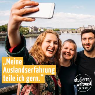 Drei lachende Studierende machen vor einer Landschaft mit Fluss ein Selfie.