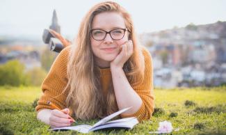 Eine versonnen lächelnde junge Frau liegt auf einer Wiese, vor sich ein Notizheft, in der Hand einen Stift. 