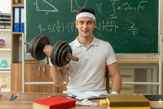 Das Bild zeigt einen fitten jungen Lehrer mit Hantel in der Hand vor einer Tafel am Lehrerpult.
