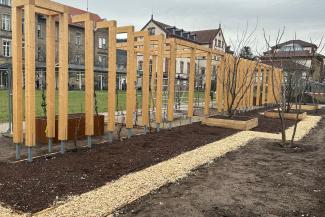 Blick auf den Klimawandelgarten in der Bauphase im Januar 2026. Das Bild zeigt Beete, Hochbeete und hölzerne Rankhilfen vor einer Grünfläche und Unigebäuden auf dem Campus Bergheim. 