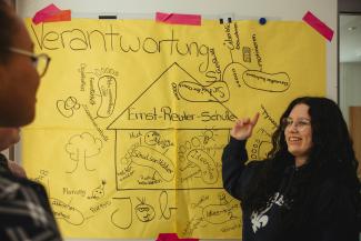 Eine junge Frau vor einem großen gelben Plakat, auf dem als Haus die Ernst-Reuter-Schule eingezeichnet ist. Die Überschrift lautet Verantwortung. Die Slkizze vermerkt in Worten und Bildern, wer wie an der Schulgemeinschaft beteiligt ist.