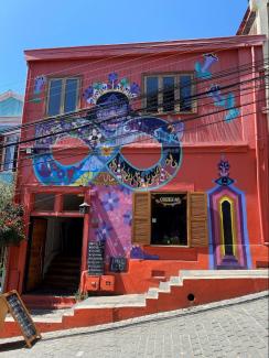 Das Bild zeigt ein rotes Haus mit buntem Mural auf einer Straße in Hanglage in Valparaiso. Der Himmel darüber ist strahlend blau.