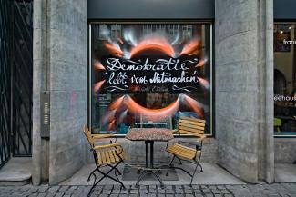 Schaufenster mit Graffiti-Schriftzug "Demokratie lebt vom Mitmachen", davor ein Tisch und 2 Stühle.