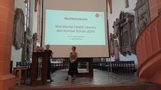 Prof. Dr. Annette Haußmann bei ihrem Vortrag in der Akademischen Mittagspause am Pult der Peterskirche. Neben ihr Rieke Deborah Koßmann. Hinter ihr eine Folie mit dem Titel ihres Vortrags.
