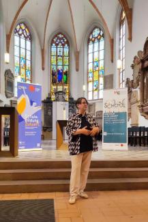 Dr. Nicole Aeschbach bei ihrem Vortrag in der Akademischen Mittagspause am Pult der Peterskirche. Hinter ihr das Roll-up zur Akademischen Mittagspause und die Buntglasfenster der Peterskirche..