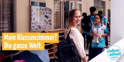 Eine junge Frau blickt im belebten Flur einer Schule über ihre Schulter; im Vordergrund gelb abgesetz der Text: „Mein Klassenzimmer? Die ganze Welt.“