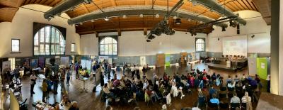 Das Panoramabild zeigt die gut besuchte Lernortmesse in der Mehrzweckhalle der PH Heidelberg, von der Empore aus fotografiert. Links im Bild Messestände, mittig Bestuhlung, rechts die Bühne bei einem der Vorträge.