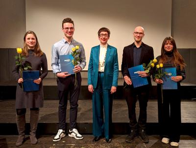 Die Preisträger:innen auf der Bühne mit Prof. Dr. Karin Vach in ihrer Mitte. In den Händen halten sie ihre Urkunde und eine gelbe Rose.