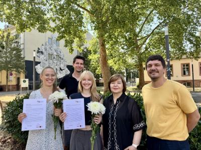 Die Absolvent:innen Julia Düchting und Laura Schweikert halten auf dem grünen Campus Bergheim ihre Zertifikate und Blumen in den Händen.  Hinter ihnen Geschäftsführer Dr. Sebastian Mahner, rechts Prof. Dr. Havva Engin und Dr. Daniel Vetter