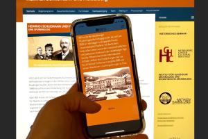 Vor dem Hintergrund der Website zur Ausstellung „Schliemann und Heidelberg“ hält eine Hand ein Smartphone, auf dessen Screen die digitale Schnitzeljagd via Actionbound zu sehen ist.