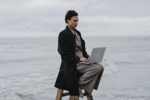 Vor winterlicher Meereskulisse sitzt eine junge Frau im schwarzen Mantel auf einer aufgeklappten Leiter in der Brandung. Auf ihrem Schoß ein aufgeklappter Laptop.