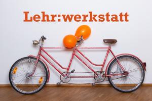 Ein orangefarbenes Tandem lehnt an einer Wand, am hinteren Lenker sind 2 orangefarbene Luftballons angebunden. Darüber der Schriftzug „Lehr:werkstatt“ in Orange.