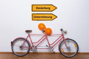 Ein orangefarbenes Tandem lehnt an einer Wand, am hinteren Lenker sind 2 orangefarbene Luftballons angebunden. Darüber der Schriftzug „Lehr:werkstatt“ in Orange, außerdem 2 Wegweiser: Auf dem einen steht „Heidelberg“, auf dem anderen „Unterstützung“. 