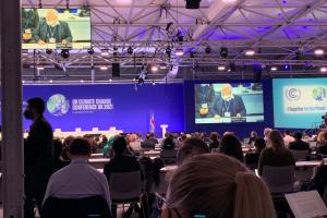 Szene auf der UN-Klimakonferenz in Glasgow 2021 (Conference of the Parties – COP26): Über die Köpfe vieler Teilnehmender hinweg sind große Screens zu sehen, die Tagungstitel und -Logo sowie einen Sprecher der EU zeigen. | © Max Jungmann
