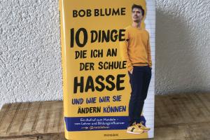 Auf einem hölzernen Tisch steht angelehnt an die Wand das Buch „10 Dinge, die ich an der Schule hasse und wie wir sie ändern können“ von Bob Blume. Die gebundene Ausgabe ist in Gelb gehalten und zeigt neben dem Titel ein Ganzkörperbild des Autors in gelbem Sweater und gelben Sneakers.