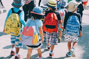 Eine von hinten abgelichtete Gruppe Grundschüler:innen mit Rucksäcken und Sonnenhüten auf dem Weg zur Schule