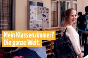 Eine junge Frau blickt im belebten Flur einer Schule über ihre Schulter; im Vordergrund gelb abgesetz der Text: „Mein Klassenzimmer? Die ganze Welt.“