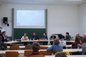 In einem Hörsaal der Neuen Universität sitzen die 6 Teilnehmenden der Podiumsdiskussion vor dem Publikum; links im Bild Dr. Florian Grafl.  