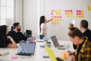 Studierende mit Laptops in einem modernen Seminarraum. Am Whiteboard eine Dozentin mit bunten Post-its.