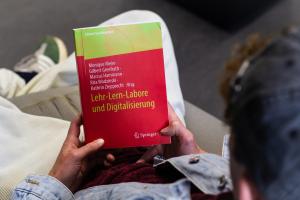 Über die Schulter eines jungen Mannes ist der Sammelband „Lehr-Lern-Labore und Digitalisierung“ zu erkennen, den er in seinen Händen hält. Das Buchcover ist rot-gelb gehalten.